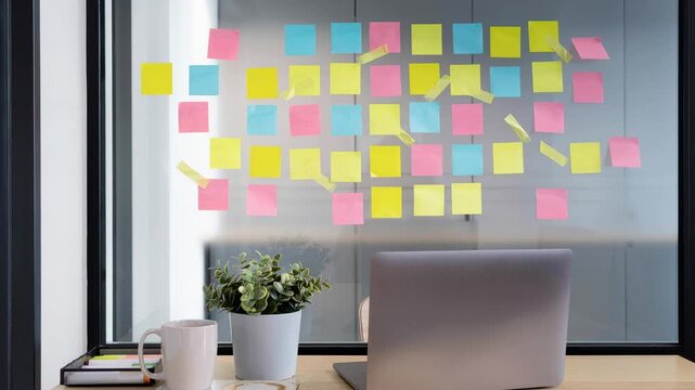 Organized colorful sticky notes on glass wall with laptop and coffee cup in modern office workspace