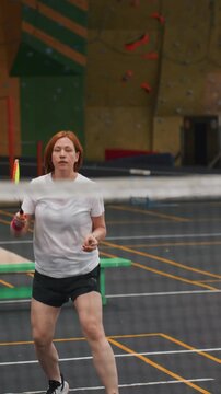 White woman playing badminton indoors, intense training session showing amateur athlete practicing footwork, serving and smashing near net, determined expression, sweatband on wrist, gym bench