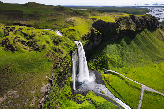 Aerial view of Seljalandsfoss waterfall cascading over a steep mossy cliff into a pool with walking paths and green meadows Rangarthing eystra, Iceland.