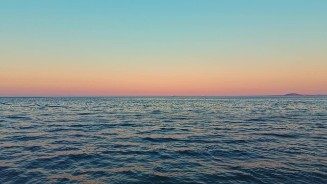 Waves move softly across the ocean as the sky changes colors from blue to orange and pink at sunset. A distant land outline is visible on the horizon at evening time.