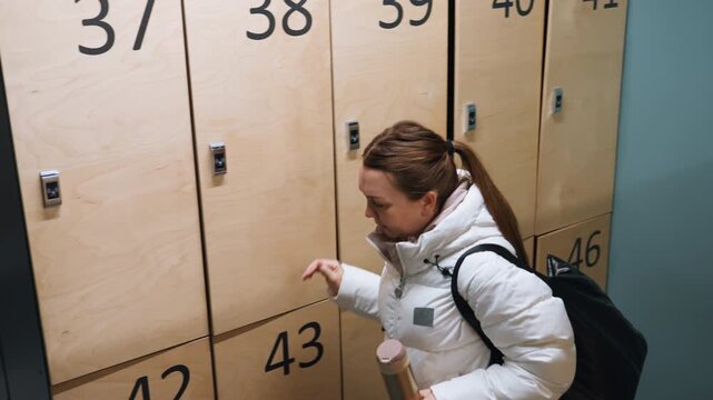 Locker room caucasian woman preparing badminton wearing white puffer jacket carrying black duffel unlocking numbered wooden locker holding reusable bottle early morning focus solitary athlete arriving