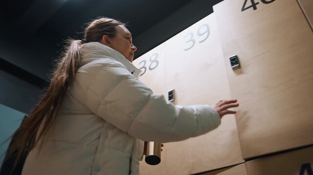 White woman at wooden lockers retrieving thermos and bag puffer jacket, long ponytail, numbered cabinets in dim modern changing room athlete grabs water, student collects books, commuter stores coat