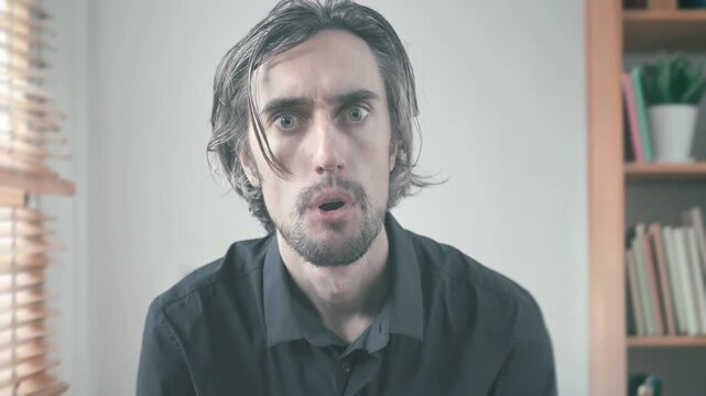 Man with Long Hair and Beard Nods Respectfully in Surprise and Sadness While Sitting Against a Wooden Blinds Background in Soft, Subdued Lighting