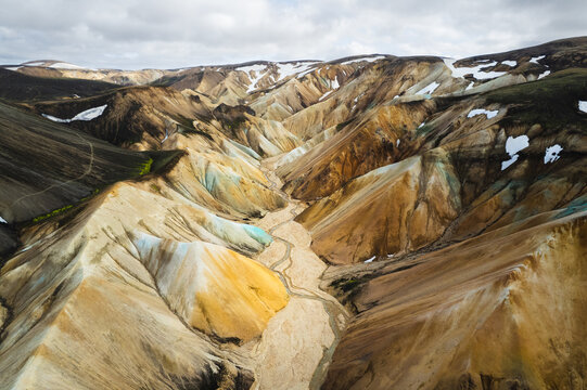 Aerial view of Landmannalaugar rhyolite mountains with colorful slopes, a winding river valley, and patches of snow under a cloudy sky Rangarthing ytra, Iceland.