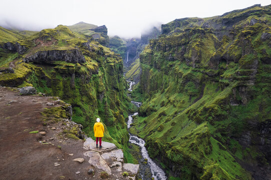 Aerial view of a person in a yellow jacket standing on a cliff edge overlooking Mulagljufur Canyon with its lush green walls and a winding river Eastern Region, Iceland.