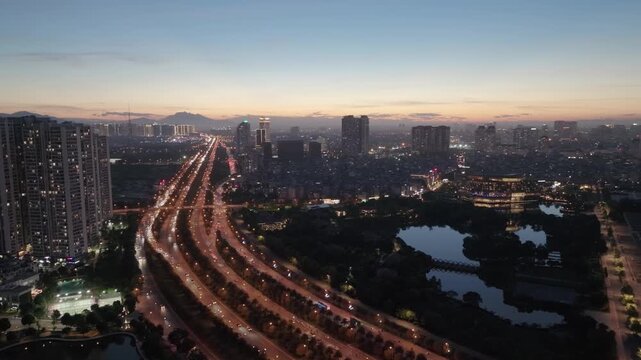 Blue hour drone aerial of Hanoi boulevard with glowing traffic trails beside lakes and modern urban skyline.