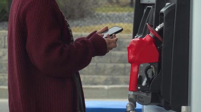 Close-up of a person in a maroon sweater lifting a red fuel nozzle to refuel at a gas station.