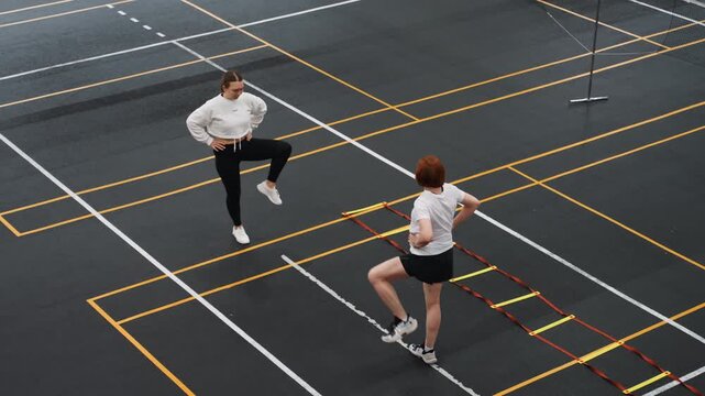 Badminton court partners executing ladder warmup, two players mirroring quick knee lifts and lateral steps on marked indoor court near net pole, high energy preparation and match focus
