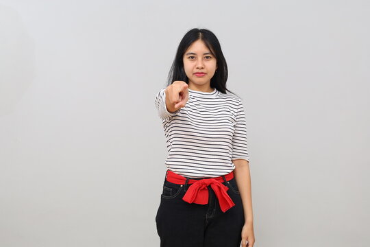 A young woman points to the camera in front of her with a commanding, angry, or accusatory expression, standing isolated against a white background.