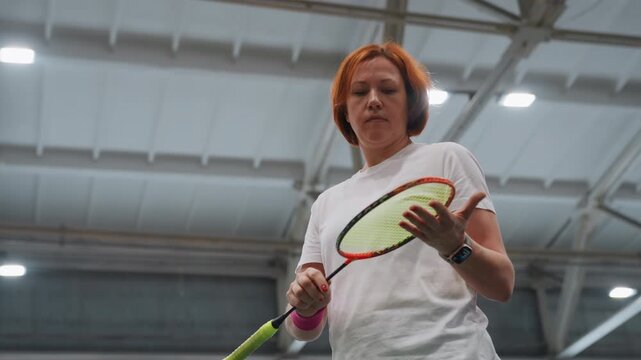 Redhaired white woman inspecting racket during indoor badminton warmup, coach inspecting grip and strings, focused expression, gymnasium lighting, slow deliberate movements, quiet preparation