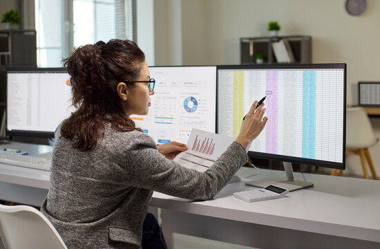 Businesswoman analyst in office reviews analysis on monitor. She points at a spreadsheet, compares charts and uses multi screen tools to verify finance files. Data fuels wise business choices.