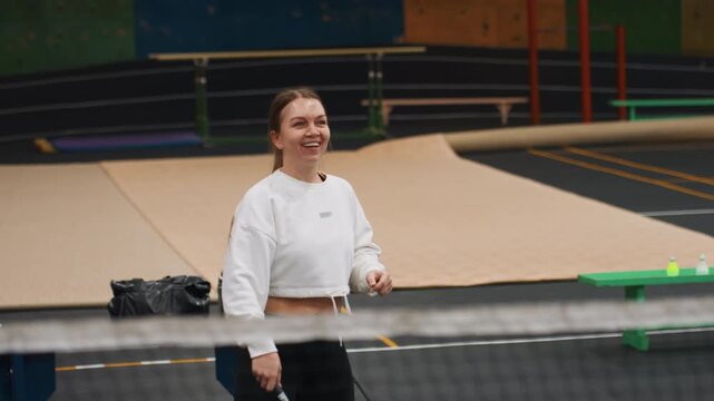 Smiling white woman playing badminton on indoor court near net, recreational player warming up with racket and shuttlecock, relaxed sweatshirt, light drills, laughter and upbeat energy, emphasis