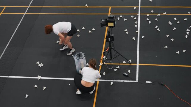 Players clean up. Team members cooperate to tidy up shuttlecocks after intense practice. Players collaborate in collecting shuttlecocks around feeder during postsession cleanup and laughter