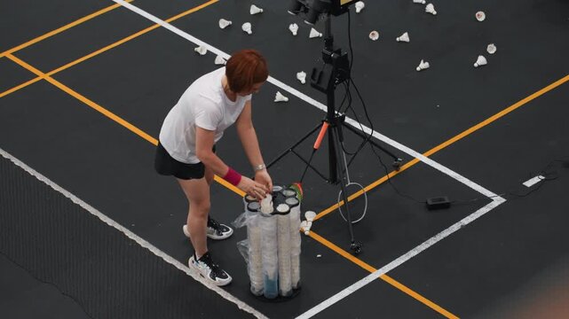 Athlete prepares shuttlecock dispenser on court, overhead shot shows scattered shuttlecocks, tripodmounted camera and training machine, concentrated warmup routine and systematic equipment arrangement