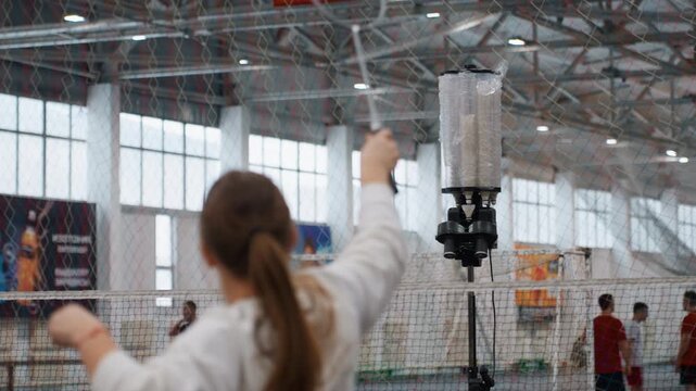 Indoor badminton practice with shuttle launcher. Teen player seen from back hitting repeated feeds from automated machine, net dividing court, coaches and teammates in background, focused technique