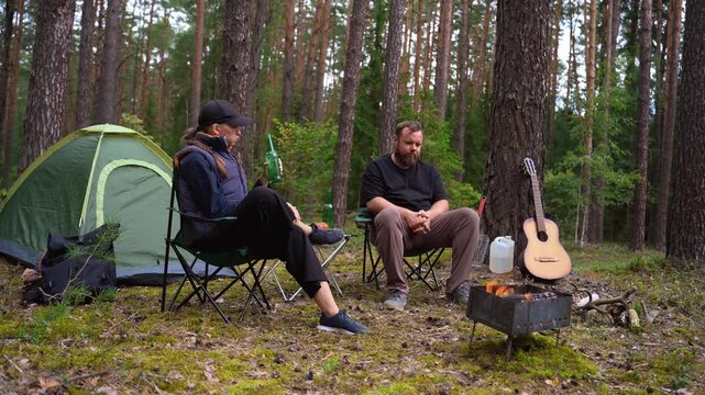 Young man and woman relaxing in camping chairs by a campfire, drinking beer, and talking in a beautiful pine forest while a guitar rests against a tree during their weekend getaway