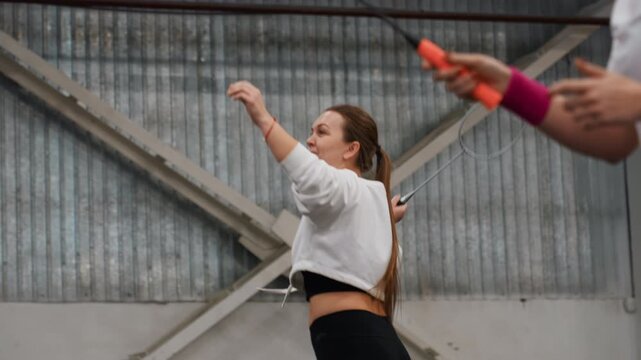 White woman practicing badminton indoors, amateur player warming up with braided hair and sweatshirt, watching shuttle flight, gymnasium metal beams, calm focused expression, slow practice swings