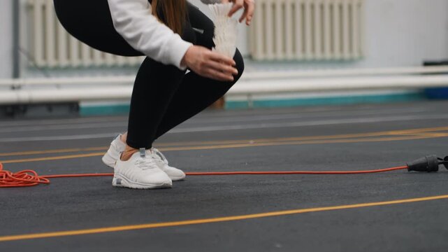 Closeup of shuttlecocks on indoor court, player crouching to arrange shuttle pile near court lines and rope, white sneakers, careful foot placement, calm warmup ritual before drill, precise hand