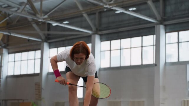 Indoor gym caucasian coach demonstrating badminton drills collecting shuttlecocks and executing repetitive serves, high ceiling windows and fluorescent lights, brisk footwork and focused posture, club