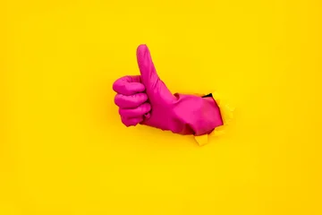 Pink cleaning glove thumbs up through torn yellow paper background © GVS