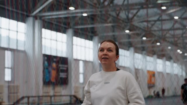 Focused caucasian woman practicing badminton indoors, wristy shots with racket, eyes tracking shuttlecock, white sweatshirt, industrial gymnasium, large windows and banners, controlled footwork