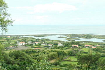 Aerial View of Azores Coastal Village and Green Landscape Description: Bird's-eye view of a small village nestled between green hills and the Atlantic Ocean on the Azores islands, showcasing the drama © Ina