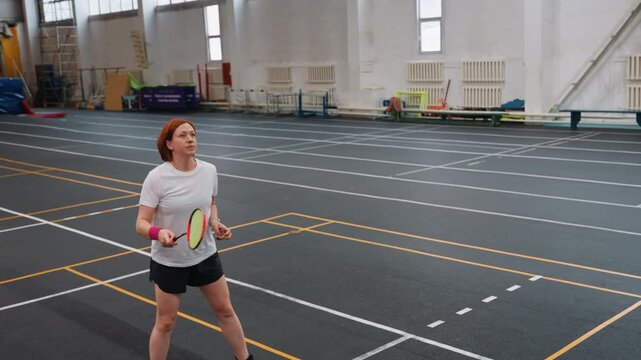 Female player practicing badminton indoors on marked court serving and tracking shuttlecock with green racket focused footwork and light training drills casual gym atmosphere with empty bleachers