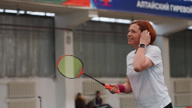 Caucasian woman playing badminton indoors smiling during casual club practice, white tshirt and pink wristband, red hair, controlled racket swings, gentle volleys and net exchanges, light gymnasium