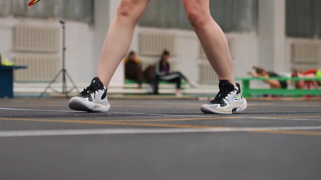 White athlete badminton footwork on indoor court, coach observing from sideline, closeup legs and sneakers executing lateral shuffle and quick steps, rubber flooring with court markings, focused