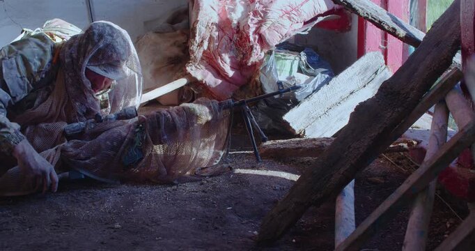 A sniper in an abandoned barn lies down and begins to take aim at the enemy. He and his rifle are camouflaged with protective cloths. Slow motion 4k footage.