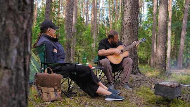 Young couple enjoying a quiet evening at a campsite where a bearded man plays an acoustic guitar for a woman while relaxing in camping chairs next to a tent and a small campfire