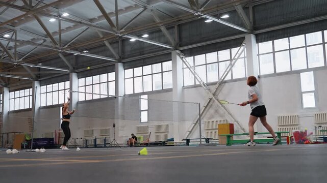 Indoor badminton court caucasian coach instructing player through technical drills, coach offering feedback while athlete practices serves and footwork, large gym hall backdrop with cones
