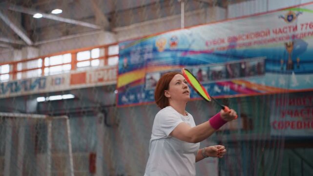 Caucasian woman executing badminton smash indoors, explosive forehand strike with wristband accent, banners and high ceiling gym backdrop, power and intensity in motion, comeback training scene