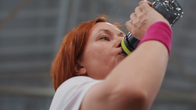 Player pauses to drink water midgame. Athletic woman takes quick water break during competition. Focused female athlete rehydrates between rallies during intense indoor badminton match