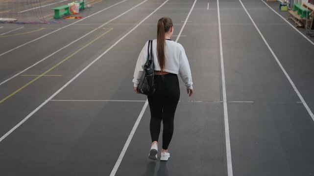 Caucasian woman walking away indoor track purposeful pace with long ponytail and gym bag slung over shoulder, heading toward courts and warmup area empty lanes and soft echoes create focused