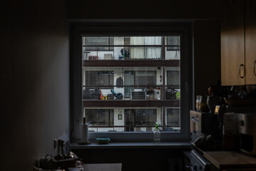 Moody view from dark kitchen interior through window to opposite apartment building with balconies, curtains and everyday objects, soft natural daylight, urban lifestyle concept, framed perspective. © Viktoria