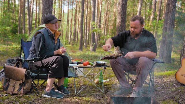 Young man and woman enjoying a relaxing picnic in a pine forest, with the man grilling meat on a barbecue while the woman watches, surrounded by camping gear and a tranquil atmosphere