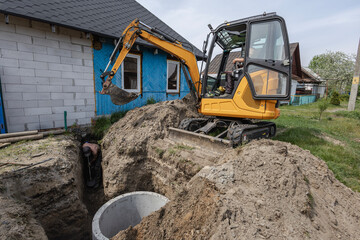An excavator digs a trench next to a house for installing a drainage system. The sky is clear and work is taking place in a rural setting © Anoo