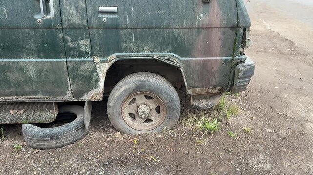 Abandoned green minibus with flat tire, rusted panels and moss-covered windows punctured spare wheels strewn on gravel parking