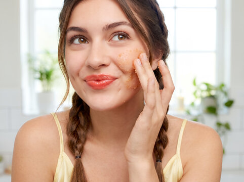 White woman applying face scrub exfoliator on cheek smiling brunette braided pigtails yellow strap top in bright bathroom plants morning skin care routine beauty cosmetic exfoliation lifestyle moment
