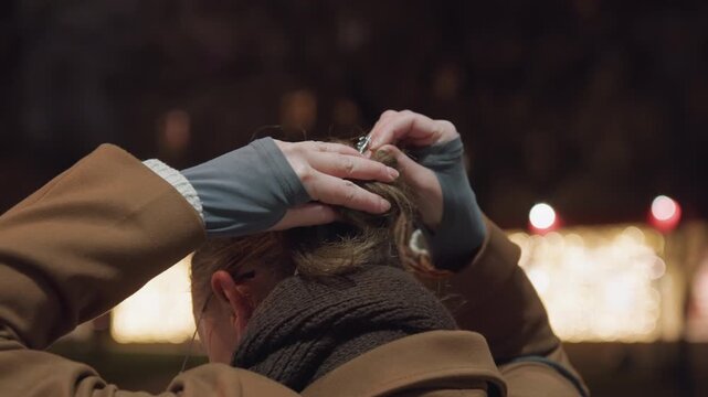 Evening park portrait of white woman adjusting wool scarf and hair bun, city bokeh lights, cold breath, thoughtful mood, glasses, brown coat, hands smoothing hair, candid rear profile.