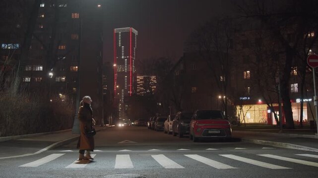 Night crosswalk under neon tower, lone pedestrian crossing zebrastriped street, parked car with glowing headlights, lit storefronts and redlit skyscraper in distance, falling snow on sidewalks,