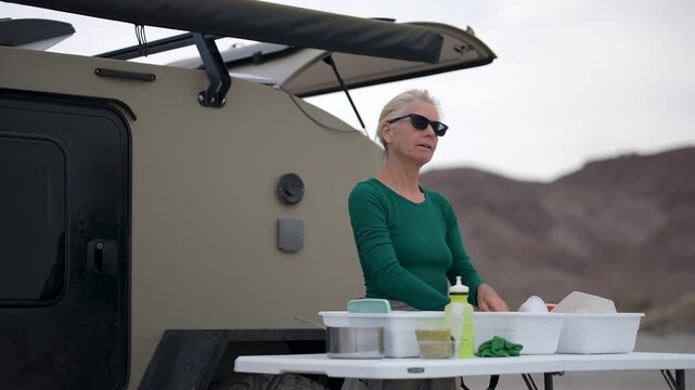 A mature woman cleans dishes at a camping setup with a teardrop trailer on a beach in Baja California, Mexico.