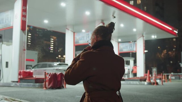 Night gas station caucasian woman in coat pausing under neon canopy, checking phone, cold breath visible, distant traffic and parking lot lights, cinematic urban solitude and quiet reflection.