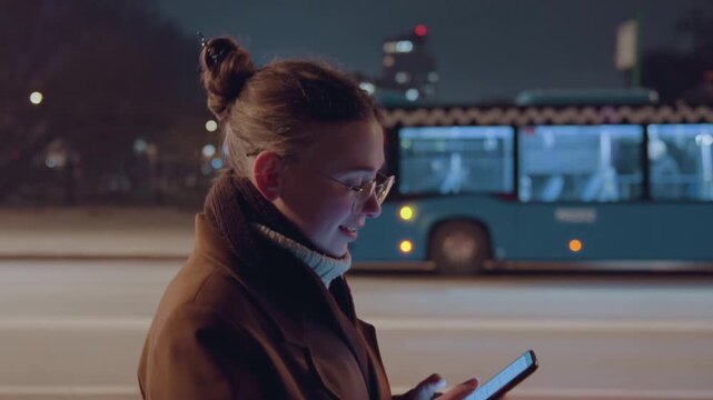 Young woman waiting at bus stop with phone, city bus passing behind, profile portrait in brown coat and scarf, checking messages and timetable, patient commuter routine, warm lamppost glow