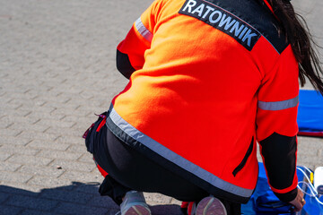 Back view close-up of a paramedic wearing a bright orange fleece jacket with "RATOWNIK" text, kneeling on a paved area with medical equipment, bright sunlight, reflective safety stripes and dark work  © Adam