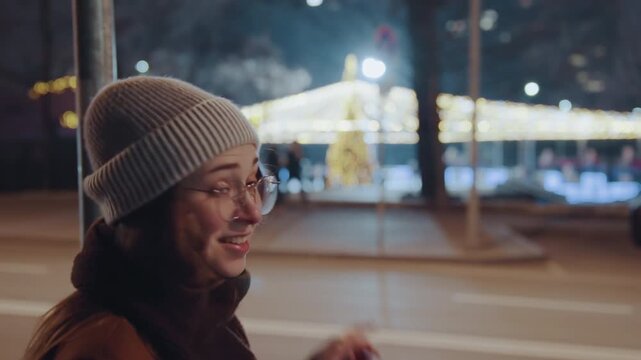 Young caucasian woman gasping at lights on city avenue, beanie and glasses, covering mouth then pointing, playful vlogger energy, festive bokeh, chilly evening walk, candid urban reaction scenes