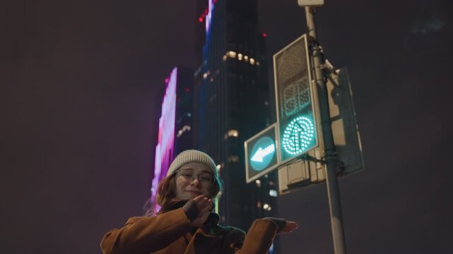 Young caucasian woman under neon sign and pedestrian signal, smiling and giving thumbs, coat and beanie, downtown highrise backdrop, content creator pose, vibrant urban night energy