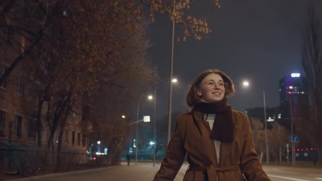 Carefree night woman with bag dancing under streetlights arms outstretched smiling face brown coat beanie empty avenue neon signs energetic motion cinematic bokeh autumn chill joyful mood