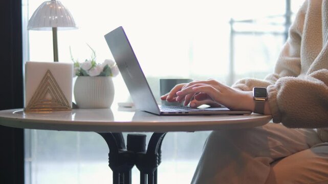 Person typing on laptop at round table. Table holds lamp, small potted plant, and candle. Background shows window with soft natural light. Person wears cozy sweater and smartwatch